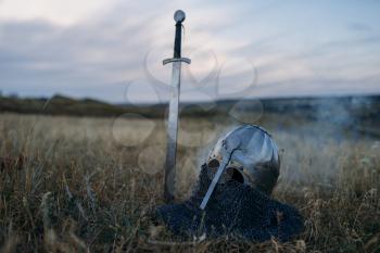 Sword stuck in the ground and metal knight helmet, nobody, great warriors symbol in the field