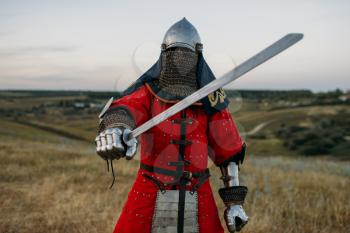Medieval knight in metal helmet holds sword, great battle. Armored ancient warrior in armour posing in the field