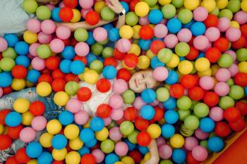 Jouful girl lying among many colorful balls in the entertainment center, top view. Female child leisures on holidays, childhood happiness, happy kids on playground