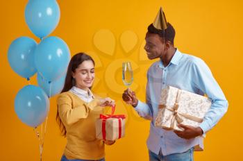 Happy love couple in caps holding glasses of beverage and gift boxes, yellow background. Pretty family party, event or birthday celebration, balloons decoration
