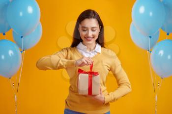 Happy woman opens gift box with red ribbons, yellow background. Pretty female person got a surprise, event or birthday celebration, balloons decoration