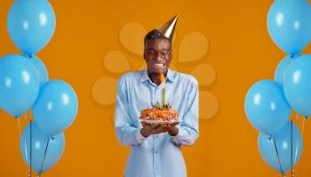 Cheerful man in cap holding birthday cake with firework, yellow background. Smiling male person got a surprise, event celebration, balloons decoration