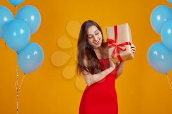 Smiling woman opens gift box with red ribbons, yellow background. Pretty female person got a surprise, event or birthday celebration, balloons decoration