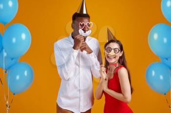 Funny love couple in caps and party masks, yellow background. Pretty family, event or birthday celebration, balloons decoration