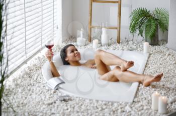 Sexy woman with glass of beverage in bath with milk. Female person in bathtub, beauty and health care in spa, wellness treathment in bathroom, pebbles and candles on background