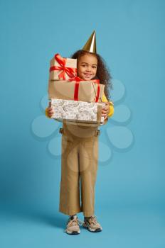 Funny little girl in cap holds birthday gift box with red ribbons, blue background. Pretty child got a surprise