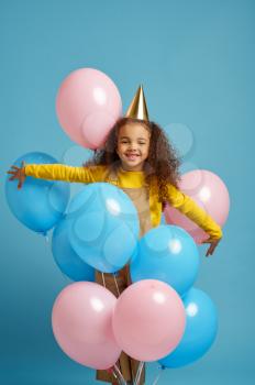 Funny little girl in cap holds a bunch of colorful balloons, blue background. Pretty child got a surprise, event or birthday party