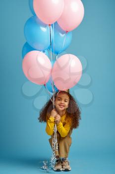 Funny little girl in cap holds a bunch of colorful balloons, blue background. Pretty child got a surprise, event or birthday party celebration