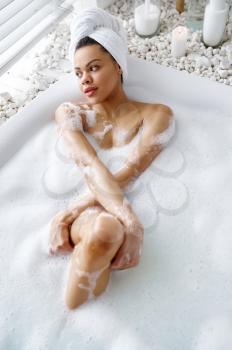 Attractive woman relax in the bath with foam. Female person in bathtub, beauty and health care in spa, wellness treathment in bathroom, pebbles and candles on background