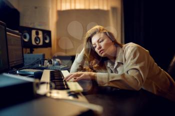 Tired female sound engineer in headphones, recording studio interior on background. Synthesizer and audio mixer, musician workplace, hard creative process