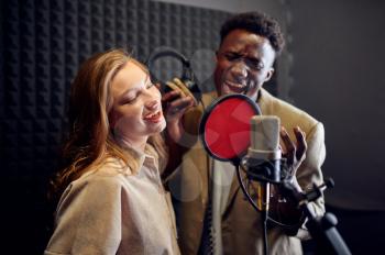 Male and female singers in headphones sings a song at micriphone, recording studio interior on background. Professional voice record, musician workplace, creative process, modern audio technology
