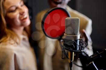 Male and female singers in headphones sings a song at micriphone, recording studio interior on background. Professional voice record, musician workplace, creative process, modern audio technology