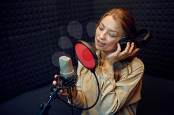 Female performer in headphones sings a song at micriphone, recording studio interior on background. Professional voice record, musician workplace, creative process, modern audio technology