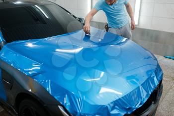 Car wrapping, man with squeegee installs protective vinyl foil or film on hood. Worker makes auto detailing. Automobile paint protection coating, professional tuning