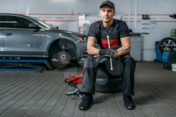 Auto mechanic with wrench sitting on wheels, tire service. Technician repairs car tyre in garage, professional automobile inspection in workshop