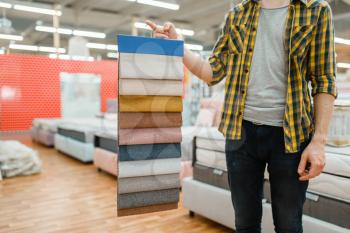 Young male person holds texture palette of mattresses in furniture store showroom. Man looking samples for bedroom in shop, husband buys goods for modern home interior