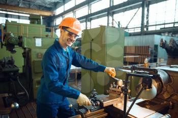 Male worker in uniform and helmet checks turning lathe on plant. Industrial production, metalwork engineering, power machines manufacturing