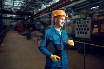 Smiling worker in uniform and safety helmet holds notebook, factory floor on background, plant. Industrial production, metalwork engineering, power machines manufacturing