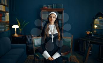 Young nun in a cassock with a cross around her neck holds a book. The sister is preparing for prayer in the monastery