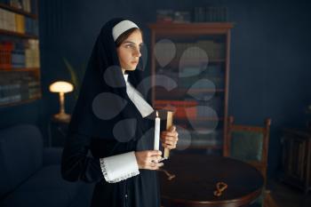 Young nun in a cassock holds a book and candle. The sister is preparing for prayer in the monastery, religion and faith, religious people