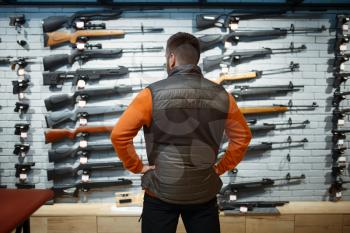 Man at showcase with rifles, back view, gun shop. Euqipment for hunters on stand in weapon store, hunting and sport shooting hobby