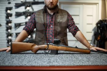 Man with rifle standing at counter in gun shop. Euqipment for hunters on stand in weapon store, hunting and sport shooting hobby