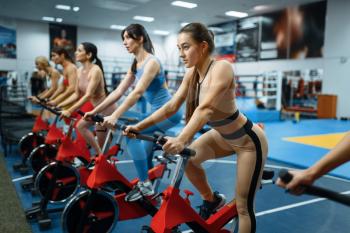 Group of women doing exercise on a stationary bike in gym. People on fitness workout in sport club, athletic girls in sportswear on training indoors