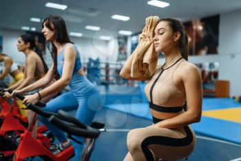 Tired woman with towel on a stationary bike in gym. People on fitness workout in sport club, athletic girls in sportswear on training indoors
