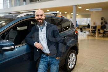Smiling man poses at automobile in car dealership. Customer in new vehicle showroom, male person buying transport, auto dealer business