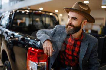 Man in hat poses at new pickup truck in car dealership. Customer in vehicle showroom, male person buying transport, auto dealer business