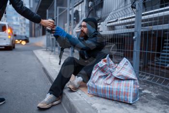 Male person gives cup of hot coffee to bearded dirty homeless on city street. Poverty is a social problem, homelessness and loneliness, alcoholism and drunk addiction, urban lonely