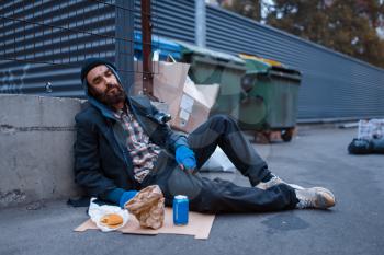 Bearded dirty beggar with food sitting at the trashcan on city street. Poverty is a social problem, homelessness and loneliness, alcoholism and drunk addiction, urban lonely