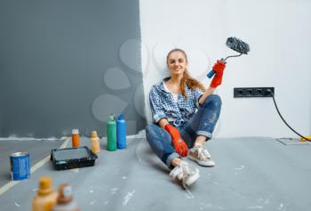 Female house painter with paint roller sitting on the floor after work. Home repair, happy woman doing appartment renovation