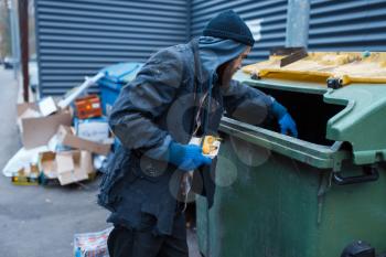 Male bearded beggar searching food in trashcan on city street. Poverty is a social problem, homelessness and loneliness, alcoholism and drunk addiction