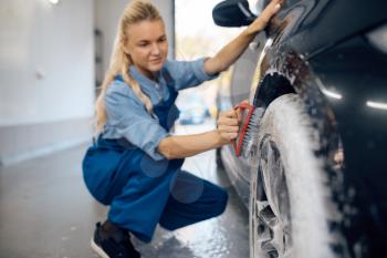 Female washer with brush in hand cleans wheel in foam, car wash. Woman washes vehicle, carwash station, car-wash business