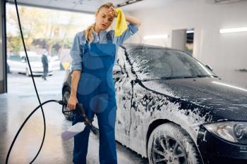 Tired female washer in uniform with foam gun in hands, car wash. Woman washes vehicle, carwash station, car-wash business