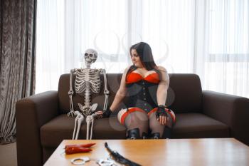 Fat depraved woman in erotic lingerie and human skeleton are sits on couch. Sexy overweight girl with big breast, corrupt large size lady