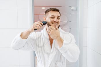 Man in bathrobe shaves his beard with an electric razor in bathroom, routine morning hygiene. Male person at the sink performs skin and body treatment procedures