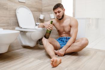 Hangover man with bottle of wine sitting on the floor near the toilet in bathroom. Hungover male person, headache and depression, bad morning