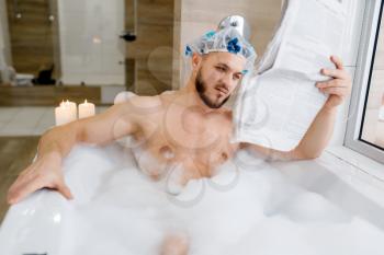 Man reads newspaper in bath with foam, morning hygiene. Male person relax in bathroom, skin and body treatments procedures