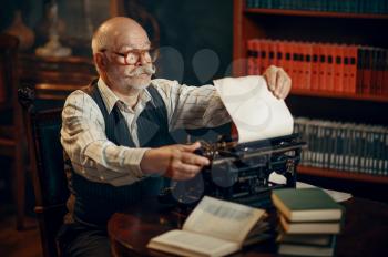 Elderly writer inserts paper into the vintage typewriter in his home office. Old man writes literature novel in room with smoke