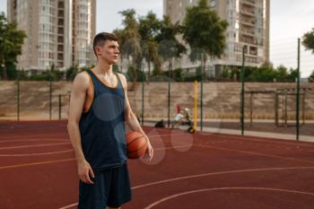 Male basketball player holds a ball on outdoor court. Male athlete in sportswear on streetball training, summer stadium