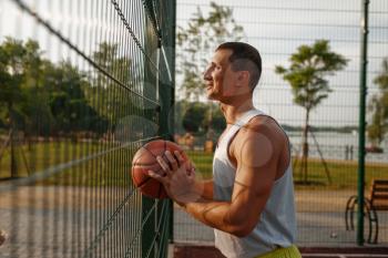 Male basketball player with ball standing at the mesh fence on outdoor court, side view. Male athlete in sportswear on streetball training, summer stadium