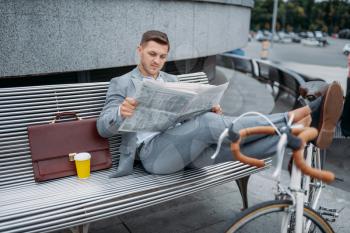 Businessman with bike reading newspaper on the bench at the office building in downtown. Business person riding on eco transport on city street, urban style