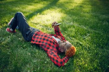 Black student with phone lying on the grass in summer park. A teenager studying and leisures outdoors