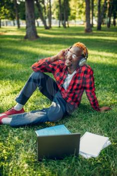 Black student in headphones sitting on the grass in summer park. A teenager studying and leisures outdoors