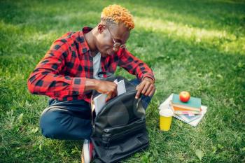 Black student with backpack sitting on the grass in summer park. A teenager studying outdoors and having lunch