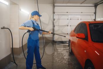 Female washer in uniform cleans the auto with high pressure gun in hands, car wash. Woman washes vehicle, carwash station, car-wash business