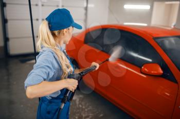 Female washer in uniform cleans the auto with high pressure gun in hands, car wash. Woman washes vehicle, carwash station, car wash business