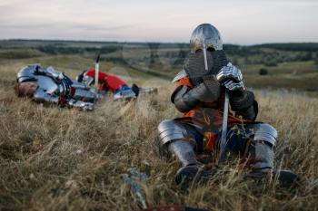 Medieval knight in armor and helmet sitting on the ground after great battle. Armored ancient warrior in armour posing in the field